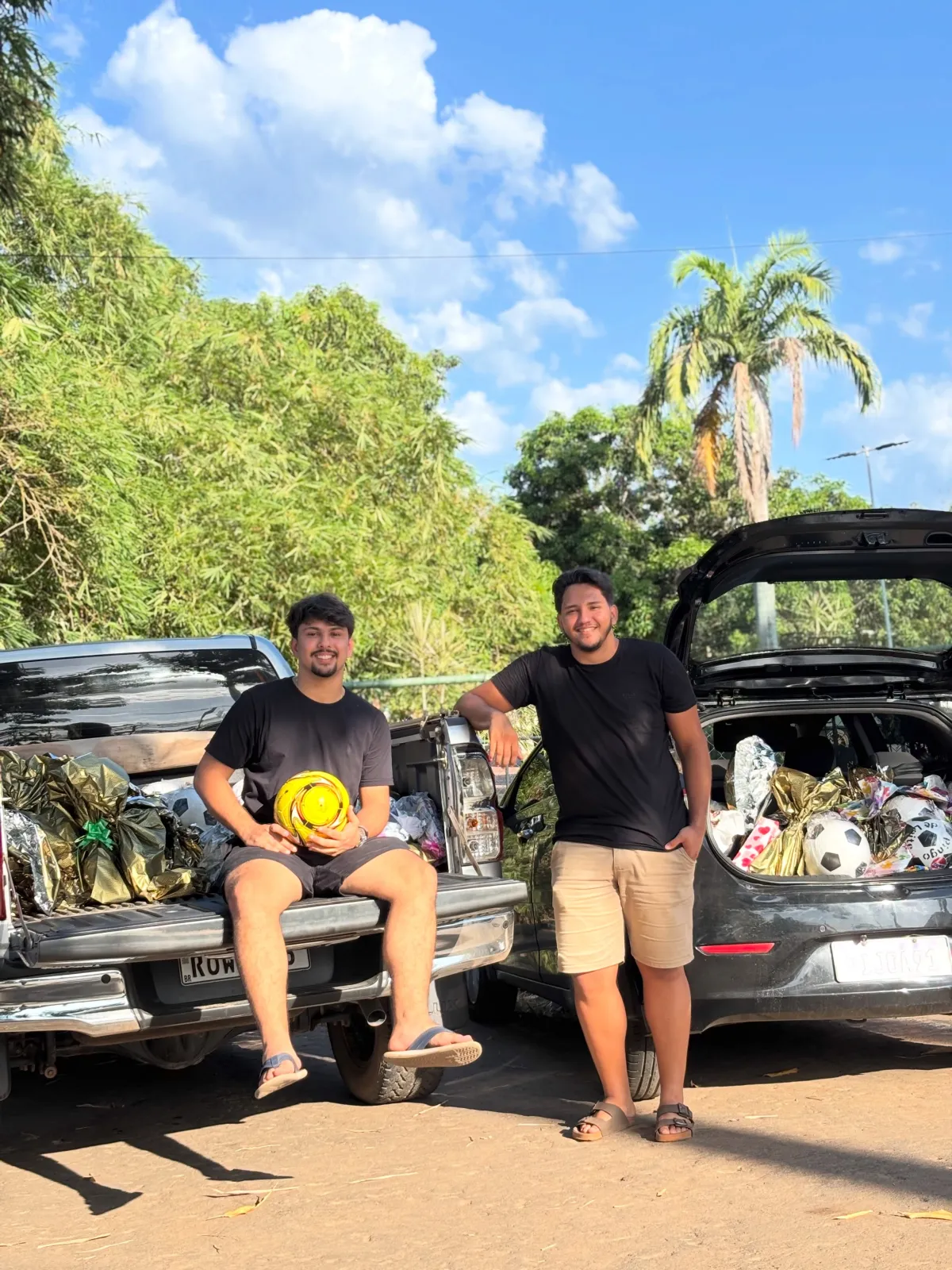 Dois jovens empresários ao lado de carros com porta-malas cheios de bolas e brinquedos para doação