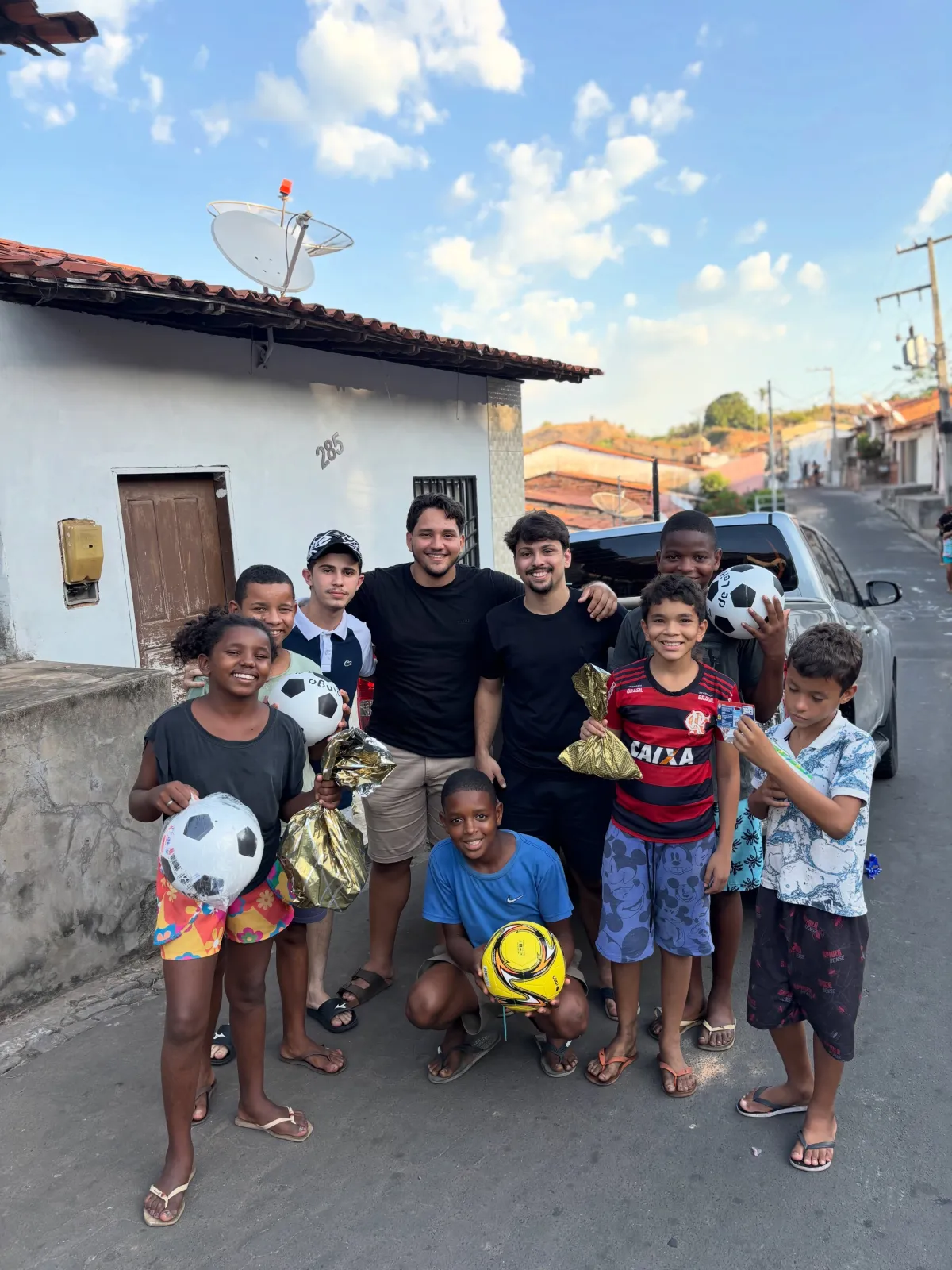 Crianças seguram bolas de futebol e doces ao lado de jovens empresários em uma rua, na ação social do Dia das Crianças